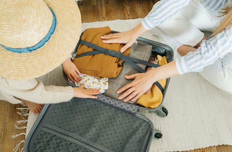 Mutter und Kind legen gemeinsam Kleidung in einen geöffneten Reisekoffer – ein Bild für sorgloses Packen, während die Hausratversicherung zuhause für Ihr Eigentum sorgt.