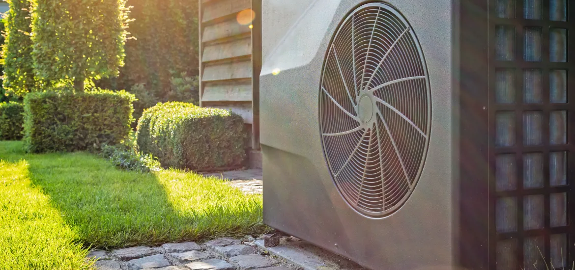 Moderne Wärmepumpe an der Außenwand eines Einfamilienhauses installiert. Im Vordergrund ein gepflegter Garten. Die Wärmepumpe symbolisiert energieeffizientes und umweltfreundliches Heizen im Eigenheim.