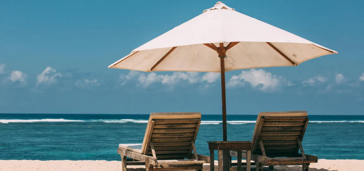 Zwei Liegestühle und ein großer Sonnenschirm am Meer – stehen für Ihren entspannten Urlaub, während Ihre Hausratversicherung während Ihrer Abwesenheit greift.